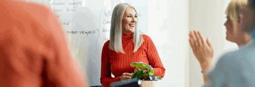 Personal perspective of mid 50s executive smiling and looking away as she receives applause from colleagues for her ideas at informal meeting.