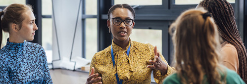 A shot of a mid-adult secondary school teacher talking with her sixth form students in class, they are wearing casual clothing and discussing what they learned in class